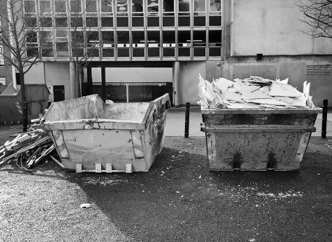Two large industrial-sized skips positioned side by side on a paved surface in front of a modern building with a grid-style facade. The left skip contains broken pieces of concrete and construction debris, with rough, uneven surfaces and a weathered, worn appearance. The right skip is filled with crumpled and torn sheets of white paper or cardboard, stacked haphazardly. Surrounding the skips, there are small scattered bits of debris and gravel on the ground. In the background, the building features large square glass panels and metal elements, with a small gate or entrance visible beneath the structure. The area appears clean but utilitarian, situated outdoors in an urban environment possibly near a commercial or industrial site, aligning with waste collection services such as those offered by Commercial Waste Cobham near [POSTCODE]. The natural lighting suggests daylight, and the overall scene indicates a typical rubbish removal setup in the Cobham area, ready for collection or disposal.