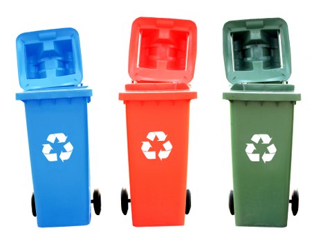 The image displays three wheeled rubbish bins aligned side by side on a plain white background. The left bin is blue with a closed lid, featuring a white recycling symbol printed on the front, and appears to be made of durable plastic with a smooth surface. The middle bin is red, also with a closed lid, bearing a white recycling symbol, and has a similar textured plastic finish. The right bin is green, positioned upright with its lid slightly open at the front, showing a similar plastic texture and finish as the other two bins. All three bins have black wheels, which are visible in the lower part of each container, and are situated in a neutral, well-lit setting that emphasizes their colours and features, consistent with waste management and rubbish collection services in the Cobham area, Surrey, serving commercial clients to facilitate recycling and waste disposal.