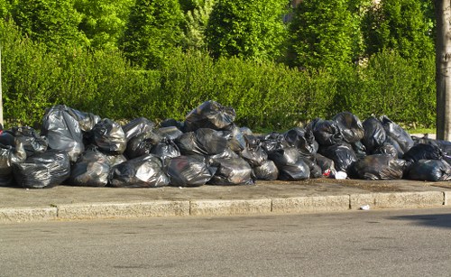 A row of large black plastic rubbish bags filled with waste is piled along the curbside on a paved street, with a concrete edge visible in the foreground. The bags are stacked haphazardly, some leaning against each other, and appear to contain household or general refuse. Behind the rubbish bags, there is a lush green hedge and a stand of trees, indicative of a suburban or residential area. The scene is lit by natural daylight, casting soft shadows, and suggests a recent collection of waste in the vicinity of Cobham within the KT11 postcode area. The image reflects the context of waste removal services, such as those provided by Commercial Waste Cobham, demonstrating the typical curbside rubbish that might be collected during commercial or domestic waste disposal operations.