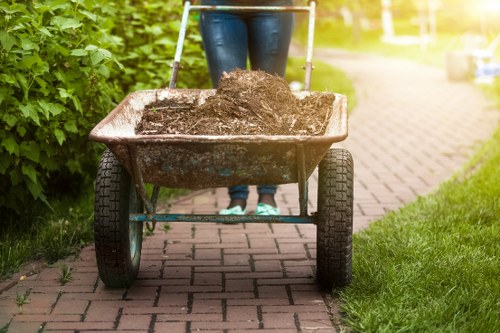 A person pushing a metal wheelbarrow along a paved pathway in a garden or outdoor area, filled with dark, loose soil. The wheelbarrow has black rubber wheels and a wooden or metal frame, positioned at the center of the image with the person's legs visible behind it. The background includes lush green bushes on the left and a lawn on the right, with sunlight illuminating the scene, suggesting a clean and tidy environment suitable for rubbish removal or garden waste collection services near Cobham. The setting appears to be part of a residential or commercial outdoor space, emphasizing the context of debris or soil clearance that Commercial Waste Cobham might handle in the local area.