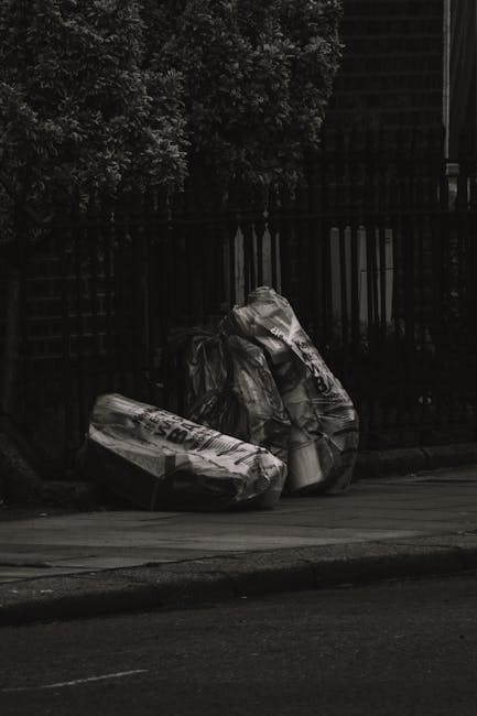 In the photograph, a large pile of mixed waste materials is situated outdoors on a paved surface, with a background of a modern industrial or commercial building featuring brick and metal siding. The 