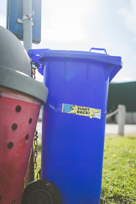 A tall, bright blue wheeled rubbish bin with a closed lid, positioned outdoors on a grassy area, close to a pink container with black holes and a grey lid. The bin features a yellow sticker with black