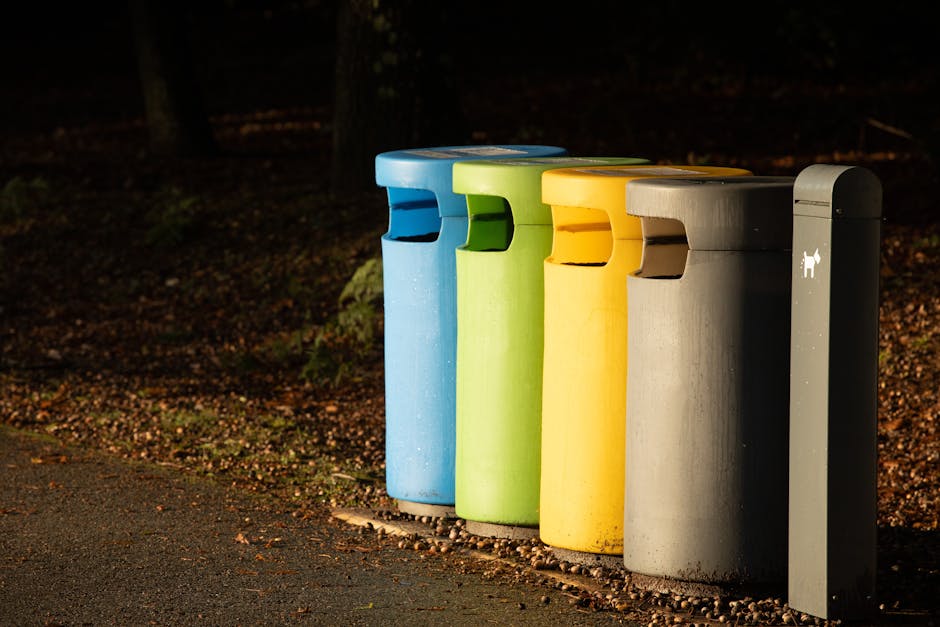 A tall, bright blue wheeled rubbish bin with a closed lid, positioned outdoors on a grassy area, close to a pink container with black holes and a grey lid. The bin features a yellow sticker with black
