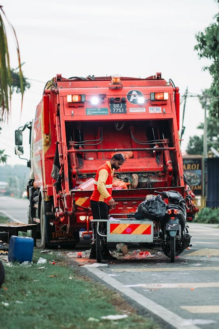 In the foreground of the image, a large overflowing bin designed for mixed paper and cardboard waste is positioned on a paved urban street. The bin’s lid is partially open, revealing a tangled colle