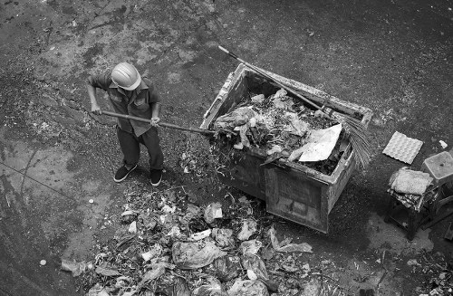 A person wearing a safety helmet and casual clothing is standing on a paved outdoor surface, using a long-handled tool to clear rubbish from a large, open-top waste skip. The skip is filled with mixed waste materials, including paper, plastic, and other debris, and is situated on a gravel or asphalt driveway. Several other waste items, such as broken pieces of cardboard or similar materials, are scattered on the ground around the skip. In the background, there are additional items, possibly tools or containers, indicating an area dedicated to waste collection or management. The environment appears to be an outdoor industrial or commercial space, with natural lighting casting shadows on the ground. This scene is typical of rubbish removal activities associated with commercial waste services, such as those offered by Commercial Waste Cobham, serving local businesses and premises in the Cobham area.